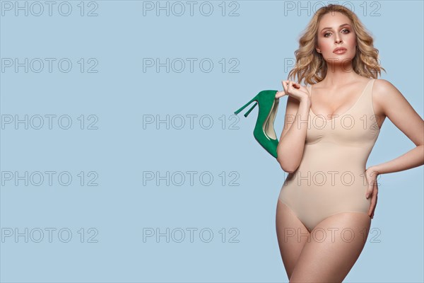Sexy beautiful blonde plus size model in lingerie bodysuit on a blue background with green shoes in her hands
