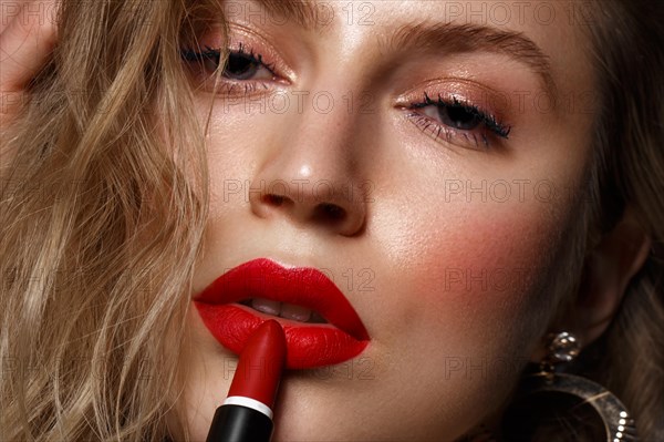 Beautiful girl with red lips and classic makeup and curls with lipstick in hand. Beauty face. Photo taken in the studio