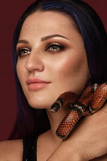 Beautiful girl brunette model with evening fashion make-up with a snake in her hands. Beauty face. Picture taken in the studio on a red background