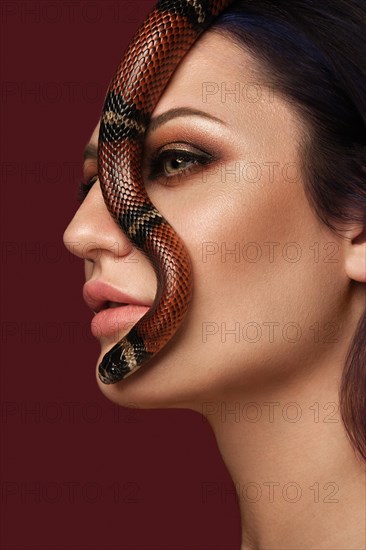 Beautiful girl brunette model with evening fashion make-up with a snake in her hands. Beauty face. Picture taken in the studio on a red background
