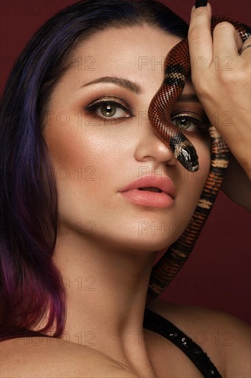 Beautiful girl brunette model with evening fashion make-up with a snake in her hands. Beauty face. Picture taken in the studio on a red background