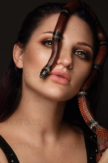 Beautiful girl brunette model with evening fashion make-up with a snake in her hands. Beauty face. Picture taken in the studio on a red background