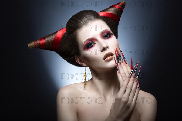 The girl in an image of the demon-tempter with long nails and haircut in the form of horns. Picture taken in the studio on a gray background