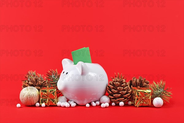 Christmas arrangement with piggy bank with green coupon surrounded by seasonal ornaments on red background