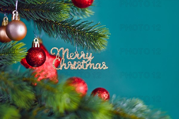 Golden Christmas tree ornament with text Merry Christmas hanging from fir tree branch with red round and star shaped baubles on teal blue background