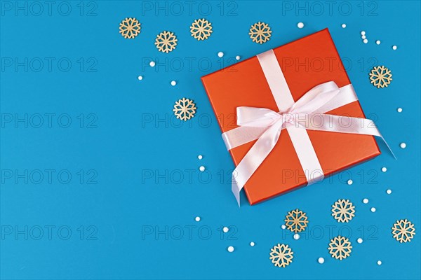 Christmas gift box with ribbon surrounded by seasonal wooden snowflake ornaments and white snowballs on side of blue background with empty copy space