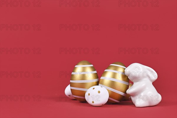 Small Easter bunny figure and golden and white easter eggs with stripes and dots on dark red background with copy space