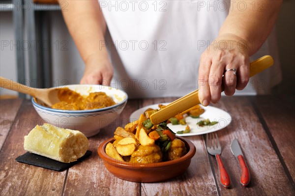 Unrecognizable woman in white apron serving potatoes and vegetables over a portion of meatballs in an earthenware dish