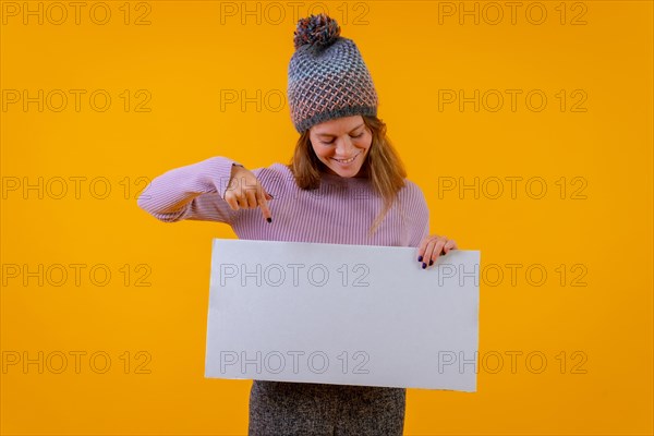 Woman in a wool cap pointing at a sign on a yellow background