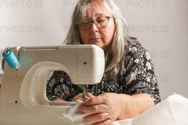 Older woman with white hair out of focus sewing a white fabric on a sewing machine