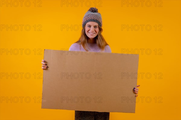 Woman in a wool cap holding a cardboard sign on a yellow background
