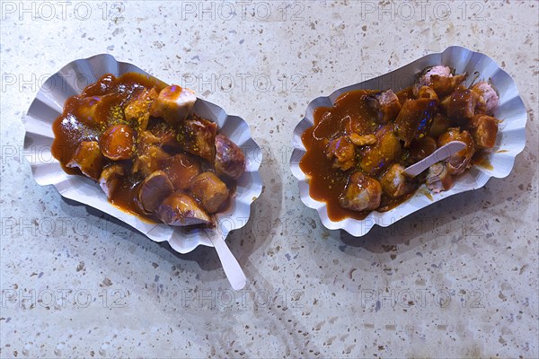 Two curry sausage portions in cardboard bowls with wooden fork at a sausage stand