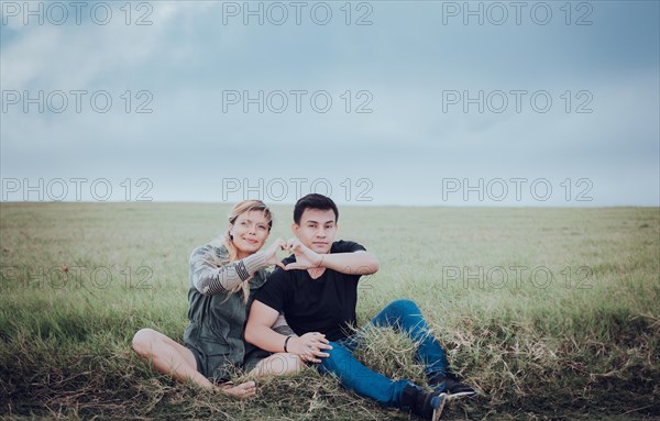 Couple sitting on the grass making a heart shape with their hands. Young couple in love in the field making heart shape. Lovers sitting in the field making a heart shape with their hands