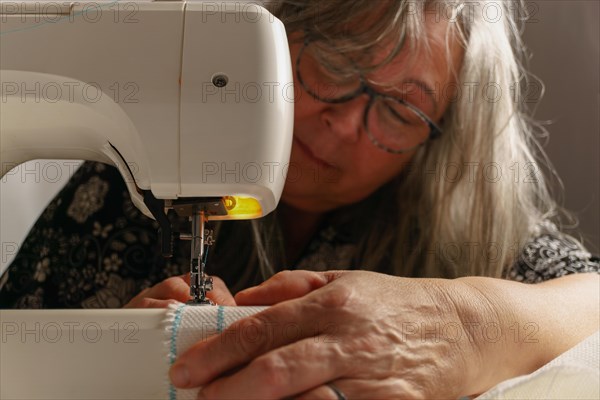 Older woman with white hair out of focus sewing a white fabric on a sewing machine