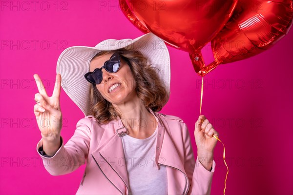 Blond caucasian woman in a white hat and sunglasses making the victory sign with her hands in a nightclub with some heart balloons