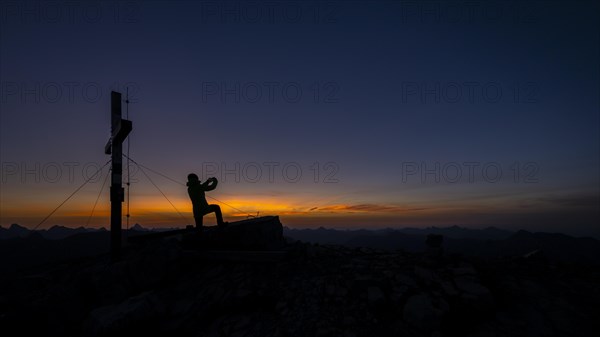 Mobile phone photographer at the summit cross of the Namloser Wetterspitze at sunset with Lechtaler Alpen