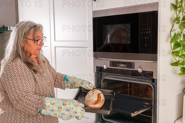 Older white-haired woman putting bread in the oven in her kitchen at home