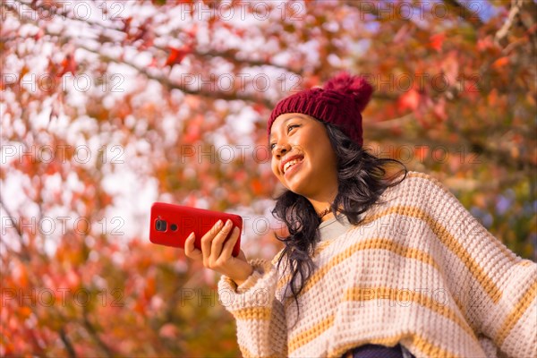 Portrait of an Asian woman in autumn with a mobile sending a voice message in a forest of red leaves