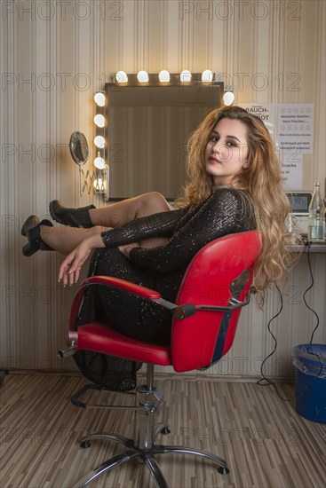 Young woman in black dress sits on a red chair in front of a dressing table