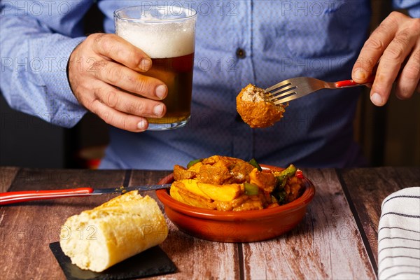 Man with beard and blue shirt eating meatballs with spaghetti in a restaurant with a glass of beer in his hand