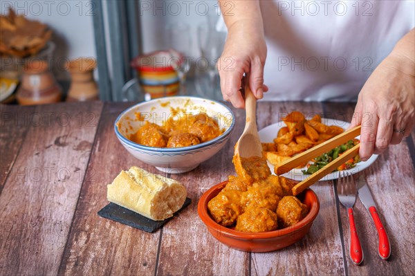 Woman in white apron serving meatballs with a wooden tong in a clay pot