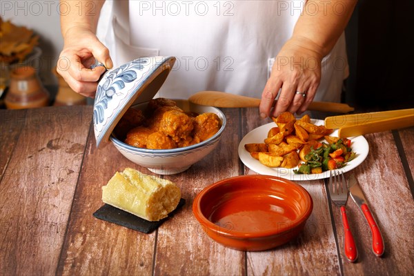 Woman in white apron serving meatballs with a wooden tong in a clay pot