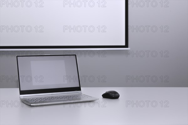 An unfolded laptop stands on a desk in front of a presentation screen