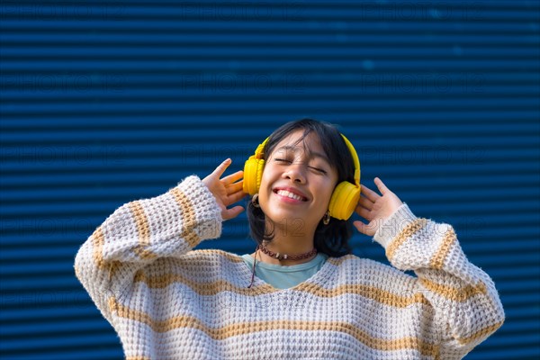 Asian girl listening to music in yellow headphones with her eyes closed on a blue college background