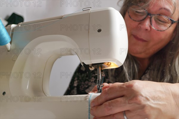 Older woman with white hair out of focus sewing a white fabric on a sewing machine