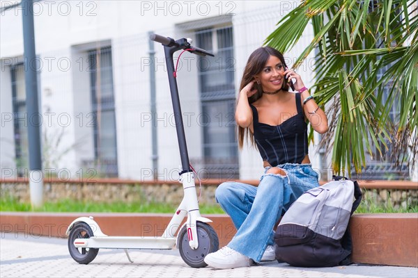 Young woman sitting in the city waiting for friends with an electric scooter talking on the phone