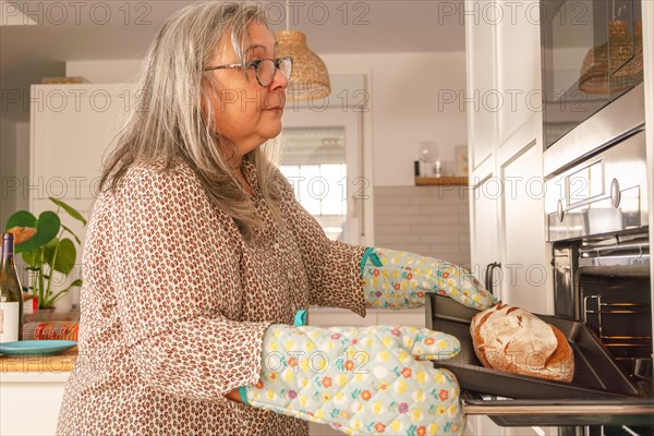 Older white-haired woman putting bread in the oven in her kitchen at home