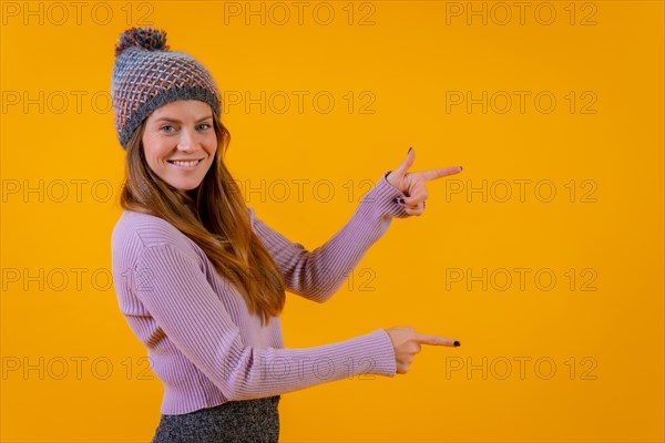Woman with wool cap on a yellow background pointing to the right smiling
