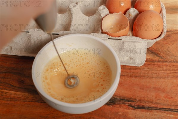 Beating an egg in a bowl with a small mixer with an egg cup at the bottom on a wooden table