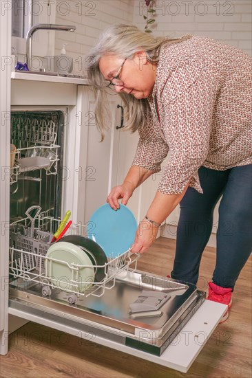 Older white-haired woman putting dishes in the dishwasher in her home kitchen