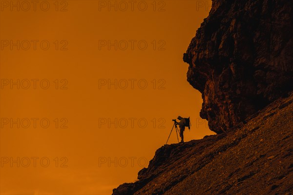 Landscape photographer in cliff face taking nature pictures of mountains with tripod at sunset in the Dolomites