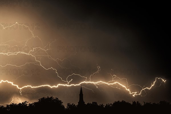 Rain falling and strokes of forked lightnings during thunderstorm at night over church tower and trees