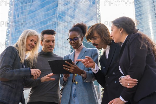 A group of smiling multi-ethnic businesspeople looking at a tablet outside the glass building