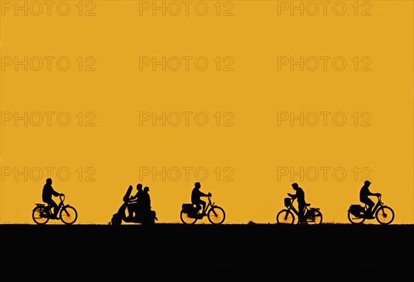 Motor scooter riding past cyclists and man on bicycle checking his smartphone silhouetted against orange sunset sky in summer