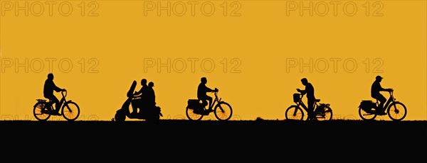 Motor scooter riding past cyclists and man on bicycle checking his smartphone silhouetted against orange sunset sky in summer