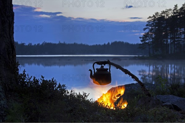 Kettle for making coffee heating over open campfire along lake at sunset