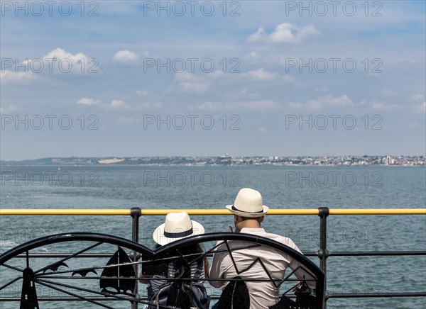 Young Asian couple sitting on a bench ovelooking the sea wearing straw hats