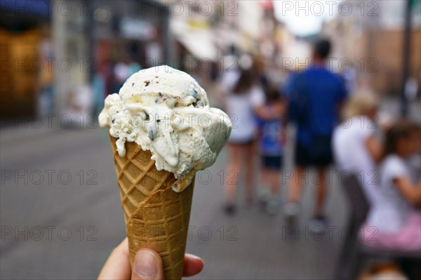 Close up of hand holding cone with vanilla with chocolate pieces ice cream with blurry city scene in background