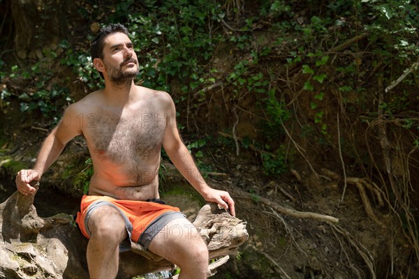 Man in swimsuit sitting on a tree trunk on a river bank