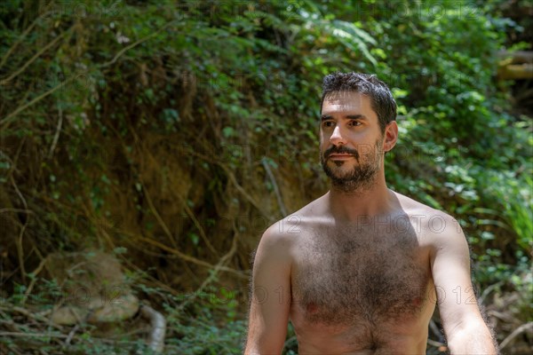 Shirtless young man sitting on the bank of a river with the forest in the background