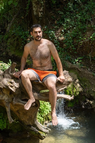 Man in swimsuit sitting on a tree trunk on a river bank