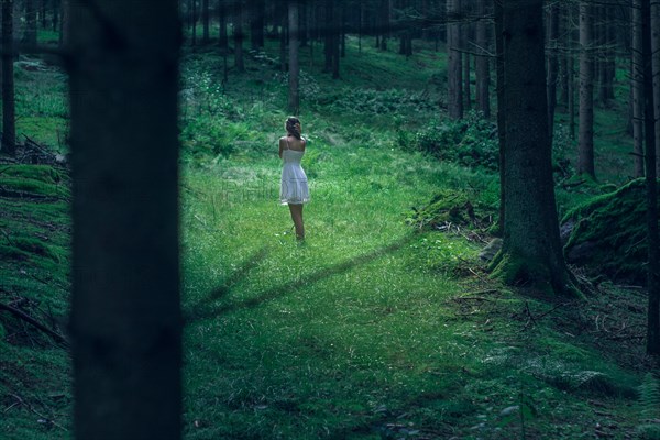 Beautiful young woman wearing elegant white dress standing in the forest with rays of sunlight beaming through the leaves of the trees