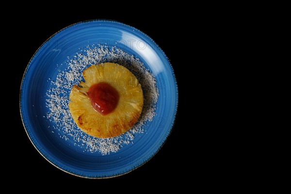 Close-up of a grilled natural pineapple with grated coconut on a blue plate on a black background