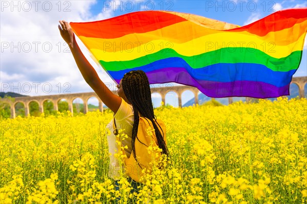 A black ethnic girl with braids holding the LGBT flag in a field of yellow flowers