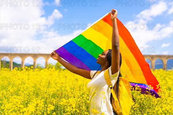 A black ethnic girl with braids holding the LGBT flag in a field of yellow flowers