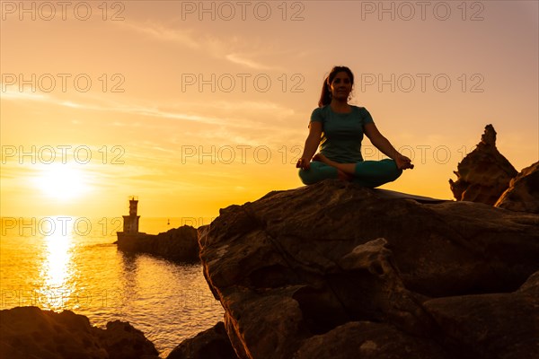 A woman doing meditation and yoga exercises at sunset next to a lighthouse in the sea
